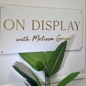 Op de afbeelding: Wit bord met gouden letters met de tekst "ON DISPLAY with Melissa Gorga".