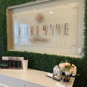 Op de afbeelding: Een witte receptiebalie met een bord waarop "PUREVIVE Medspa" staat in zilveren letters op een transparante acryl achtergrond. Het bord is gemonteerd op een muur bedekt met kunstmatig groen gebladerte.