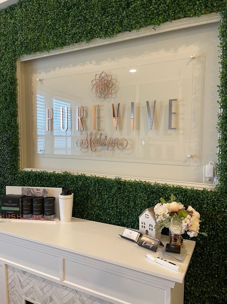 Op de afbeelding: Een witte receptiebalie met een bord waarop "PUREVIVE Medspa" staat in zilveren letters op een transparante acryl achtergrond. Het bord is gemonteerd op een muur bedekt met kunstmatig groen gebladerte.