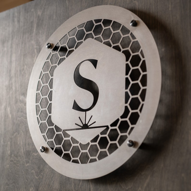 May include: A silver metal sign with a honeycomb pattern and the letter "S" in the center. The sign is mounted on a wooden wall.