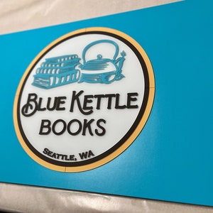 Puede incluir: Un logotipo circular azul y amarillo con un contorno negro. El logotipo presenta una pila de libros y una tetera azul con un asa negra. El texto "Blue Kettle Books" est&aacute; escrito en letras cursivas negras. Debajo del texto est&aacute; "Seattle, WA" en letras negras.