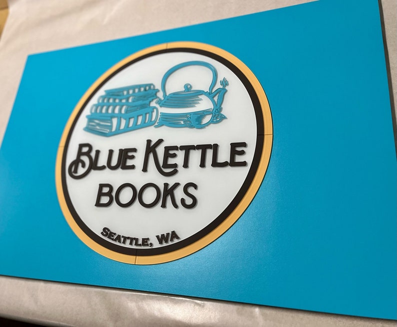 Puede incluir: Un logotipo circular azul y amarillo con un contorno negro. El logotipo presenta una pila de libros y una tetera azul con un asa negra. El texto "Blue Kettle Books" est&aacute; escrito en letras cursivas negras. Debajo del texto est&aacute; "Seattle, WA" en letras negras.
