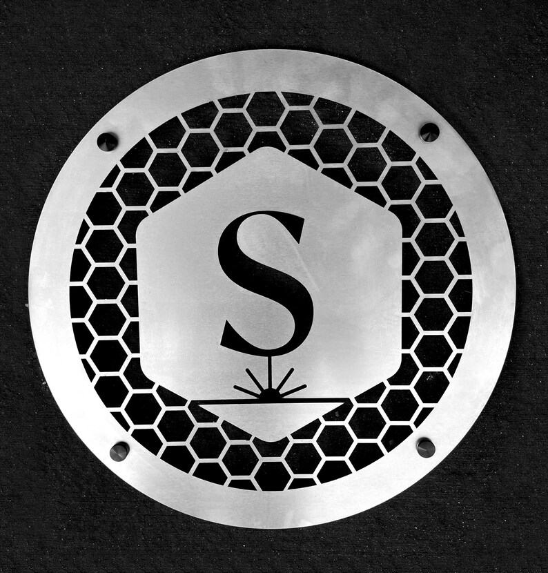 May include: A silver metal sign with a honeycomb pattern and the letter "S" in the center. The sign has two small screws on the bottom and top.