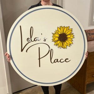 Puede incluir: Un letrero circular blanco con borde azul, que presenta el texto "Lola's Place" en escritura cursiva marr&oacute;n y un girasol amarillo con centro marr&oacute;n.