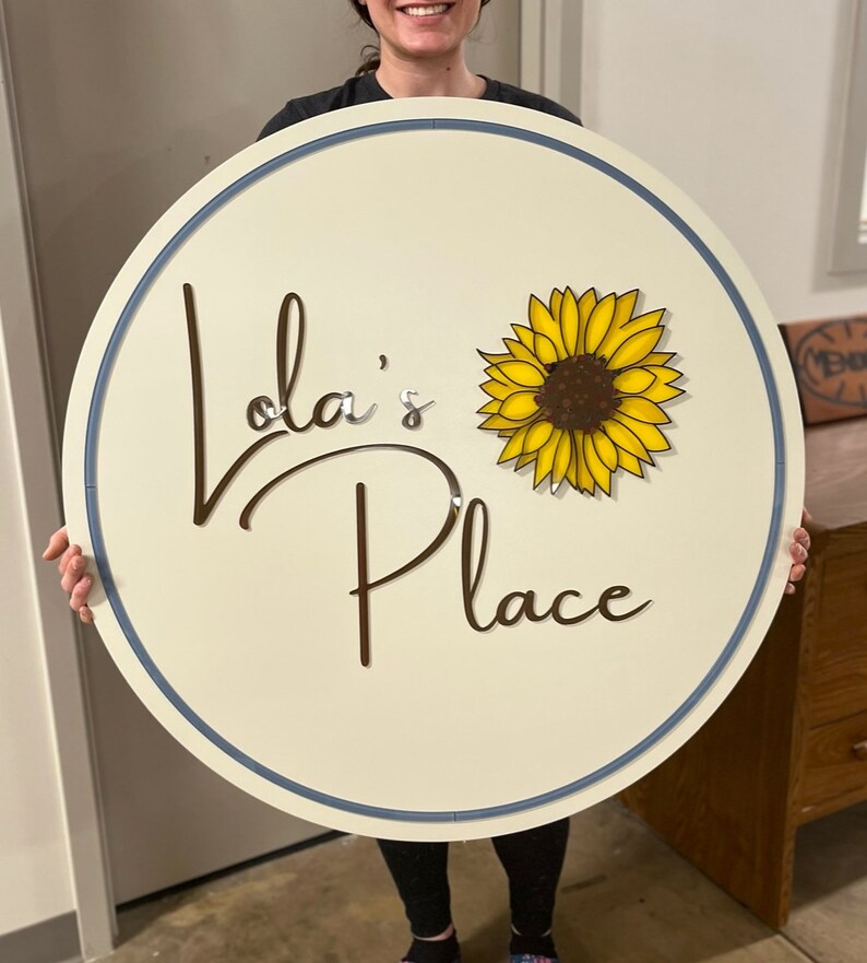 Puede incluir: Un letrero circular blanco con borde azul, que presenta el texto "Lola's Place" en escritura cursiva marr&oacute;n y un girasol amarillo con centro marr&oacute;n.