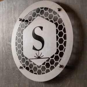 May include: A silver metal sign with a honeycomb pattern and the letter "S" in the center. The sign is attached to a wooden surface with screws.