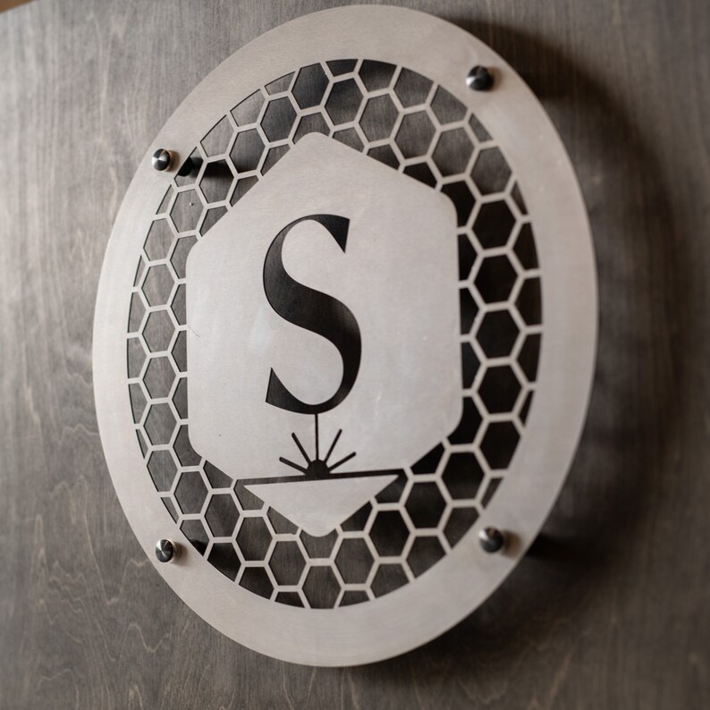 May include: A silver metal sign with a honeycomb pattern and the letter "S" in the center. The sign is attached to a wooden surface with screws.