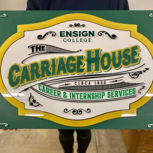 Puede incluir: Un letrero verde y amarillo con el texto "The Carriage House" en un &oacute;valo blanco. El texto "Ensign College" y "Circa 1855" tambi&eacute;n est&aacute;n en el letrero. El letrero dice "Career & Internship Services".
