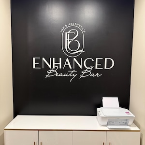 May include: A black wall featuring a white logo with the text Ink & Aesthetics, Enhanced Beauty Bar. Below the wall is a white cabinet with gold handles and a white printer on top. The logo includes a stylized letter B inside a thin white frame.