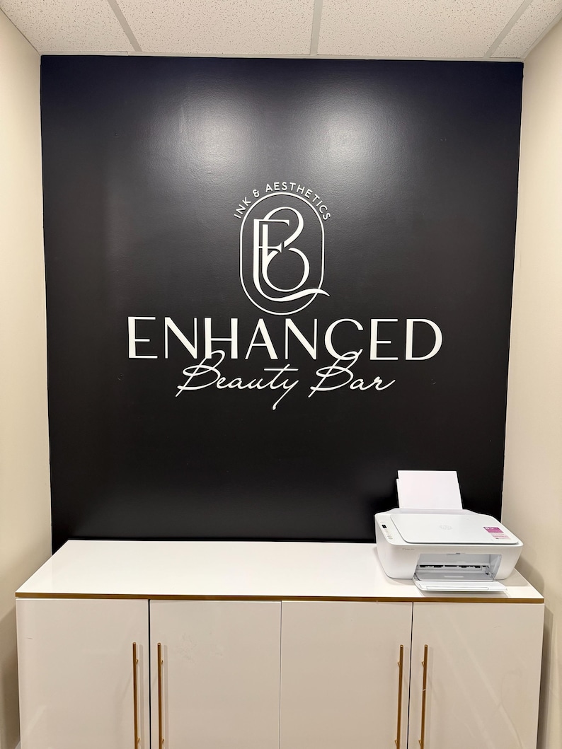 May include: A black wall featuring a white logo with the text Ink & Aesthetics, Enhanced Beauty Bar. Below the wall is a white cabinet with gold handles and a white printer on top. The logo includes a stylized letter B inside a thin white frame.