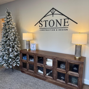 May include: A long wooden cabinet with six glass-paneled doors sits below a wall sign reading STONE CONSTRUCTION & DESIGN. Two lamps with cylindrical bases and white shades flank the cabinet, and a flocked Christmas tree stands to the left.