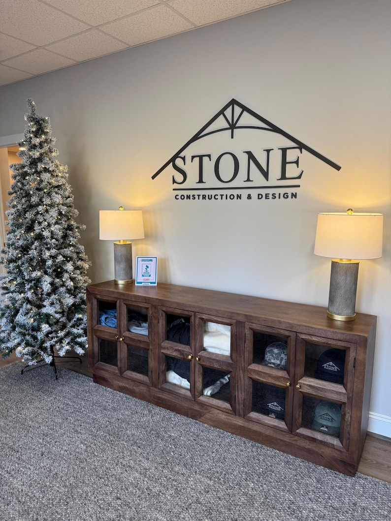 May include: A long wooden cabinet with six glass-paneled doors sits below a wall sign reading STONE CONSTRUCTION & DESIGN. Two lamps with cylindrical bases and white shades flank the cabinet, and a flocked Christmas tree stands to the left.