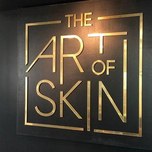 May include: A gold sign with the text "THE ART OF SKIN" in a black square frame on a black background.