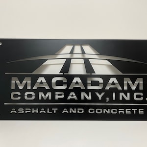 May include: Black metal sign with a silver cut-out design of a road and the text "Macadam Company, Inc. Asphalt and Concrete".