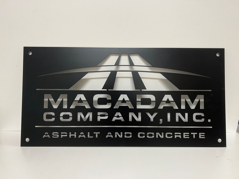May include: Black metal sign with a silver cut-out design of a road and the text "Macadam Company, Inc. Asphalt and Concrete".