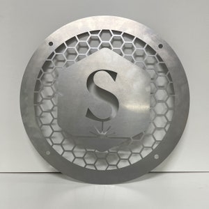 May include: A silver metal round sign with a honeycomb pattern and the letter "S" in the center. The letter is inside a hexagon shape with a small design underneath.