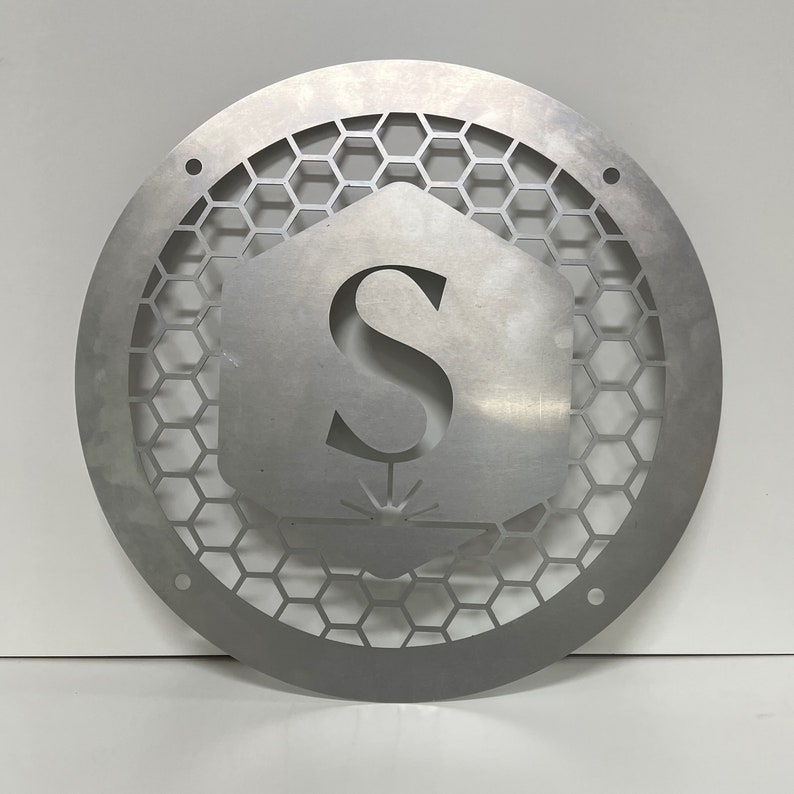 May include: A silver metal round sign with a honeycomb pattern and the letter "S" in the center. The letter is inside a hexagon shape with a small design underneath.