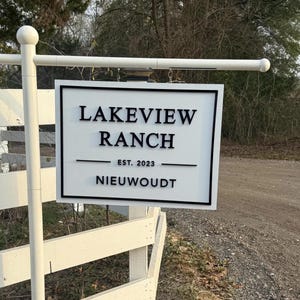 May include: A rectangular white sign with a black border hanging from a white post. The sign features black raised lettering that reads "LAKEVIEW RANCH", "EST. 2023", and "NIEUWOUDT" centered on the surface with a horizontal line separating the text.