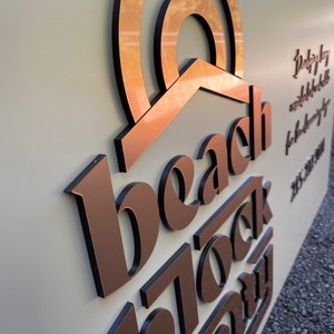 Op de afbeelding: Een koperkleurig bord met de tekst "beach block company" in zwarte, reli&euml;fletters. Het bord heeft een gestileerd zonontwerp boven de tekst.