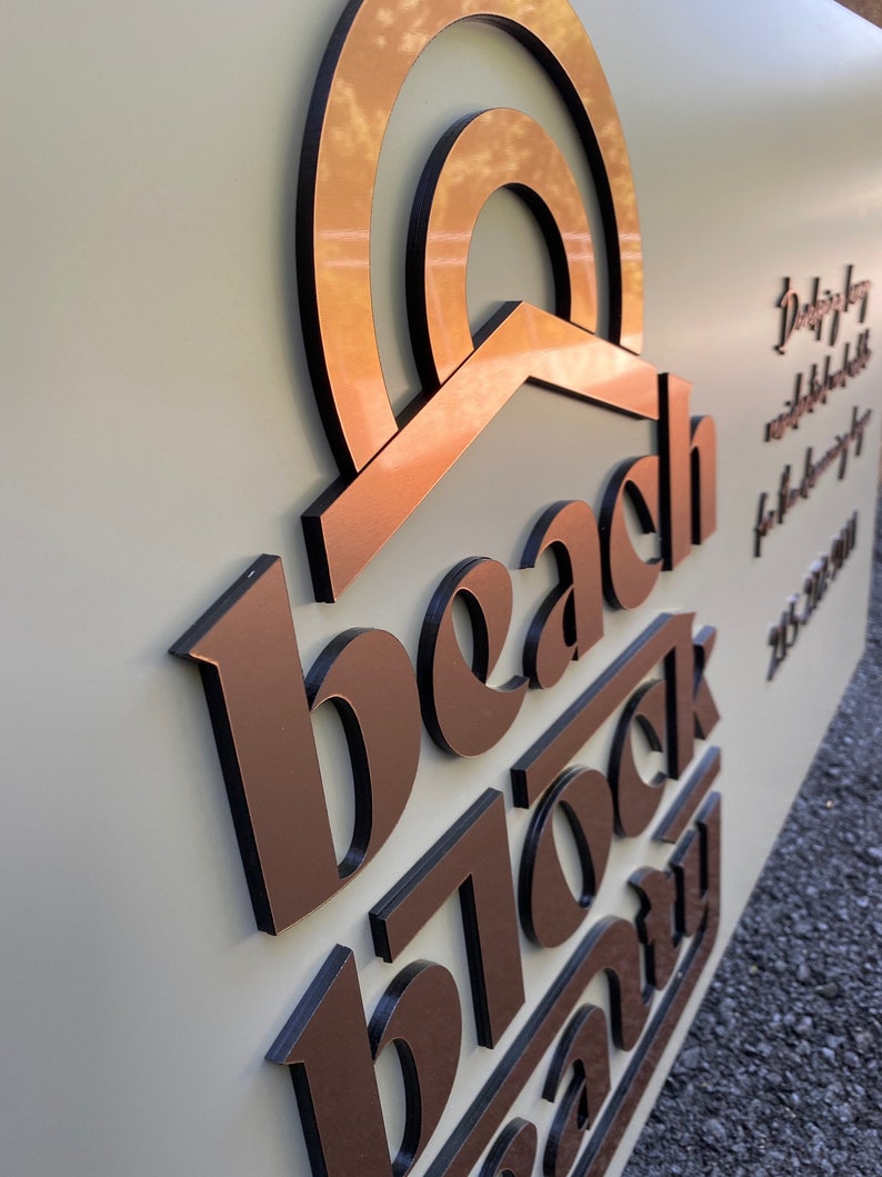 Op de afbeelding: Een koperkleurig bord met de tekst "beach block company" in zwarte, reli&euml;fletters. Het bord heeft een gestileerd zonontwerp boven de tekst.