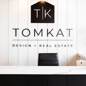 May include: A reception area featuring a white vertical paneled wall with a black logo sign reading TK TOMKAT DESIGN + REAL ESTATE. Three gold cylindrical wall lights are mounted above the sign. A black office chair sits behind a white marble counter.