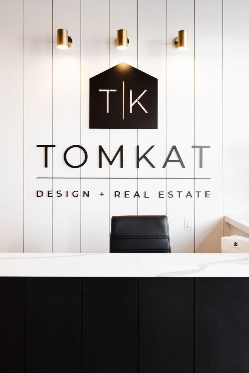 May include: A reception area featuring a white vertical paneled wall with a black logo sign reading TK TOMKAT DESIGN + REAL ESTATE. Three gold cylindrical wall lights are mounted above the sign. A black office chair sits behind a white marble counter.