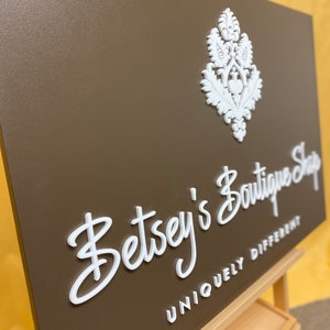 Puede incluir: Un letrero marr&oacute;n con letras blancas en relieve que dice "Betsey's Boutique Shop Uniquely Different". El letrero tiene un dise&ntilde;o decorativo blanco en la esquina superior derecha.