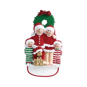 May include: A Christmas ornament featuring a family of four in red and white pajamas with a dog. The family is standing in front of a green wreath with a red bow. The ornament is white and has a blank space for personalization.