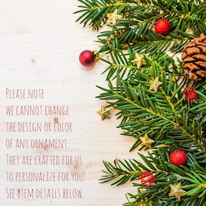 May include: A close-up of a wooden surface with a green pine branch with red and gold ornaments. The text "PLEASE NOTE WE CANNOT CHANGE THE DESIGN OR COLOR OF ANY ORNAMENT. THEY ARE CRAFTED FOR US TO PERSONALIZE FOR YOU! SEE ITEM DETAILS BELOW." is written in red.