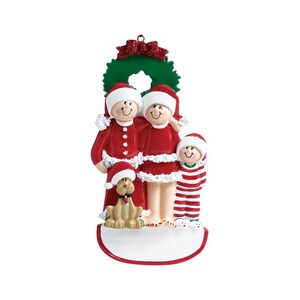 May include: A Christmas ornament featuring a family of three in red and white pajamas, standing in front of a green wreath. A small dog is standing in front of them. The ornament has a blank space for personalization.