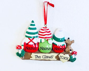 May include: A Christmas ornament featuring three gnomes with red, green, and white striped hats. The gnomes are standing on a wooden log with the text "The Claus' 20xx" and the names "Name" on each gnome.