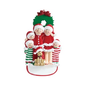 May include: A Christmas ornament featuring a family of five in red and white pajamas with a dog. The family is standing in front of a green wreath with a red bow. The ornament has a blank space for personalization.