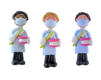 May include: Three small figurines of dentists wearing blue scrubs and face masks, holding toothbrushes and dentures. The dentists have different skin tones: black, white, and brown.