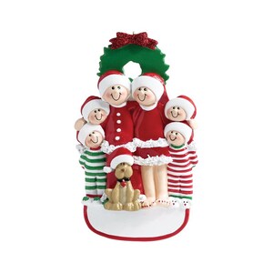 May include: A Christmas ornament featuring a family of five in red and white pajamas with a dog. The family is standing in front of a green wreath with red berries. The ornament has a blank space for personalization.