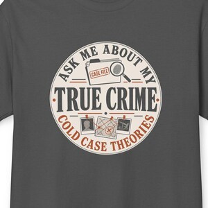 May include: A charcoal gray short-sleeve t-shirt featuring a circular graphic on the chest. The design includes a case file folder, a magnifying glass, and photos with the text: ASK ME ABOUT MY TRUE CRIME COLD CASE THEORIES.