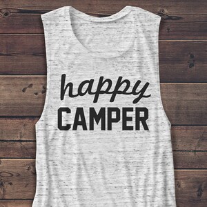 May include: A sleeveless white and gray marbled tank top with the words "happy CAMPER" printed in black. The top has a scoop neck and a curved hemline. The shirt is made of a soft, lightweight material.