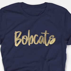 May include: A navy blue t-shirt featuring the word "Bobcats" in gold script. The t-shirt is folded and knotted at the bottom, with the sleeves rolled up.