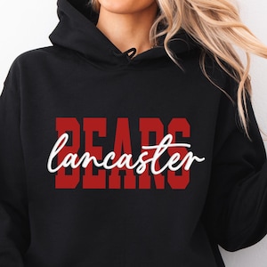 May include: Black hooded sweatshirt with the word "BEARS" in red block letters and "Lancaster" in white script. The sweatshirt has a front pocket and a drawstring hood. The model is wearing light blue jeans.