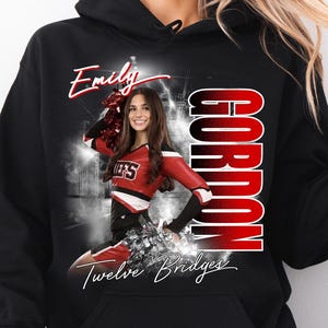 May include: Black hooded sweatshirt featuring a graphic of a cheerleader in a red and white uniform. The sweatshirt has the name "Emily" in white script, the name "Gordon" in red block letters, and "Twelve Bridges" in script.
