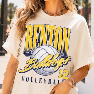 Pu&ograve; includere: Una t-shirt a maniche corte color bianco sporco con una stampa grafica che riporta il testo BENTON BULLDOGS 12 VOLLEYBALL in blu e giallo, centrato attorno a una grande illustrazione di una pallavolo. La maglia ha un girocollo e vestibilit&agrave; ampia.