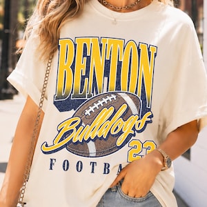 May include: An off-white short-sleeve t-shirt featuring a graphic print with the text BENTON BULLDOGS 23 and an illustration of an American football. The shirt is paired with light blue denim shorts that have a frayed hemline.
