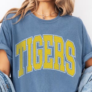 Op de afbeelding: Blauw T-shirt met het woord "TIGERS" in gele blokletters met een witte omlijning. Het shirt heeft opgerolde mouwen. Een spijkerjasje is over de schouders gedrapeerd.