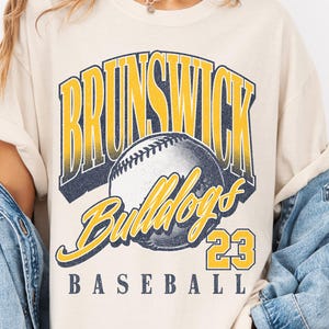 May include: A cream-colored short-sleeve t-shirt featuring a graphic print with the text BRUNSWICK in yellow and blue, a baseball illustration, the word Bulldogs in yellow script, the number 23, and the word BASEBALL in blue text.