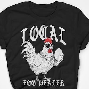 Op de afbeelding: Zwart T-shirt met een witte cartoon-haan die een zonnebril en een gouden ketting draagt. De haan houdt een ei vast. De tekst "LOCAL EGG DEALER" staat onder de haan gedrukt.