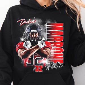 May include: Black hooded sweatshirt featuring a football player graphic. The graphic includes the name "Declan Marrone" and the number "32 Wildcats" in red and white. The player is wearing a helmet and crossed arms.