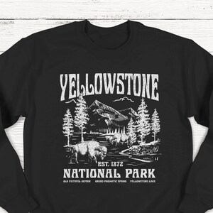 May include: Black sweatshirt with a white graphic design of a mountain landscape with trees and a bison. The text "Yellowstone National Park" is printed in white with the words "Est. 1872" and "Old Faithful, Geyser, Grand Prismatic Spring, Yellowstone Lake" printed below.