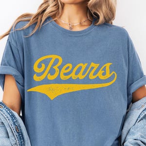 May include: Blue t-shirt with the word "Bears" in yellow script. The shirt has rolled-up sleeves and a vintage look. A denim jacket is draped over the shoulders.