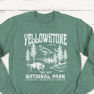 May include: Green sweatshirt with a white graphic design of a mountain landscape with trees and a bison. The text "Yellowstone National Park" is printed on the sweatshirt, along with "Est. 1872" and the names of some of the park's attractions.