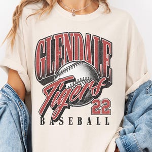 May include: A cream-colored short-sleeve t-shirt featuring a graphic print with the text GLENDALE TIGERS 22 BASEBALL. The design includes a central baseball illustration with red and black lettering, layered over a light blue denim jacket.