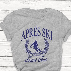 May include: Gray t-shirt with a blue graphic of a skier in a wreath with the text "Apr&egrave;s Ski Resort Social Club".